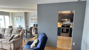 A modern living room with a sofa, a blue armchair, and a coffee table. A dining area with chairs and a table is visible in the background. The kitchen area is partially visible with a stove and oven. - Le Littoral (Magog)