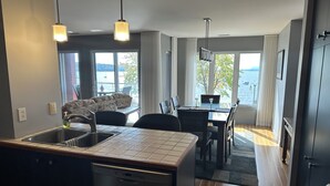 A modern kitchen with a built-in dishwasher, a dining area with a table and chairs, and large windows offering a view of the sea. - Le Littoral (Magog)