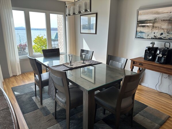 A modern dining room with a glass table, six chairs, and a view of the water. - Le Littoral (Magog)