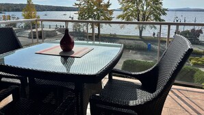 A balcony with a glass table and wicker chairs, overlooking a lake with boats and trees. - Le Littoral (Magog)