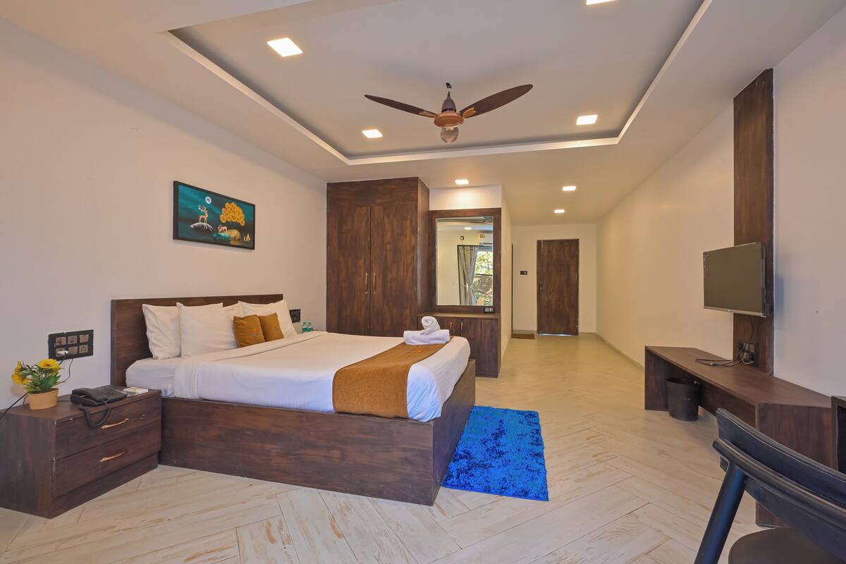 A modern hotel room with a large bed, wooden wardrobe, ceiling fan, and a TV on a wooden stand.
