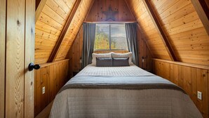 Une chambre confortable située sous les toits, avec un plafond en pente en bois, un grand lit recouvert d’une courtepointe et une fenêtre agrémentée de rideaux.