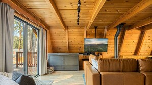 L’intérieur chaleureux d’un chalet avec un plafond en bois, un canapé, un téléviseur et une porte-fenêtre coulissante donnant sur une terrasse extérieure.