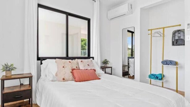 A bedroom with a large window, a bed with white bedding and multiple pillows, a bedside table with a plant, and a wall-mounted drying rack with clothes.