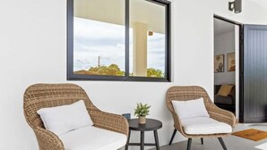 Two wicker chairs with white cushions, a small round table with a plant, and a large window with a view of a building and sky.