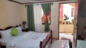 Una habitación de hotel con una cama, una mesita de noche, una lámpara, un ventanal con cortinas y vistas a un balcón con plantas en macetas.