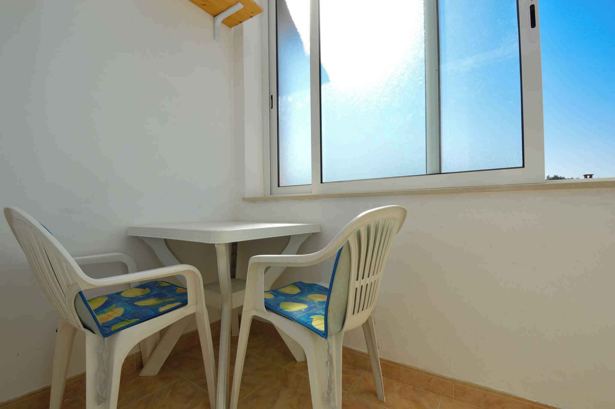 Una habitación pequeña con una mesa blanca y dos sillas con cojines azules, una ventana con vista al cielo y un suelo de baldosas.