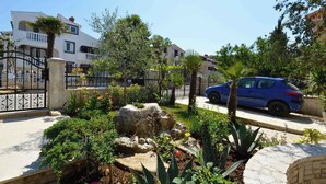 A residential area with a blue car parked on the street, a white building with balconies, and a garden with various plants and a rock centerpiece. - Ferienwohnung mit Balkon (Medulin)