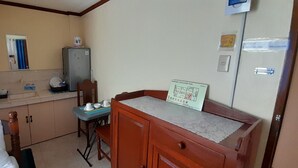Une petite cuisine équipée d’un réfrigérateur, d’une petite table et d’une chaise. On y trouve un meuble avec un tiroir et un buffet en bois recouvert d’une toile de dentelle. Un panneau est visible sur le mur.