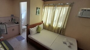 Une chambre d’hôtel compacte comprenant un coin cuisine, un lit simple avec des oreillers, une fenêtre avec des rideaux et une petite salle de bain visible à travers une porte ouverte.