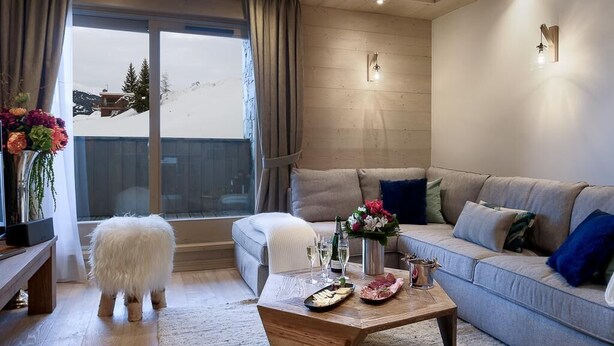 A cozy living room with a sofa, a coffee table with food and drinks, a fluffy white dog, and a view of a snowy mountain through a large window. - Wifi, 200m from resort center, 1st floor, view mountain, sauna, balcony, parking, tv, 91m² (Courchevel)