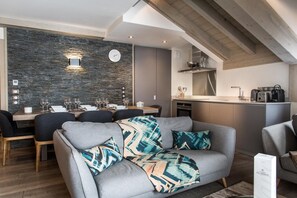 A modern living room with a grey sofa, a dining area with a stone wall, and a kitchen with a wooden ceiling.