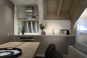 A modern kitchen with a stainless steel range hood, a built-in oven, and a coffee maker. There is a dining table with chairs and a vase with flowers on the counter.