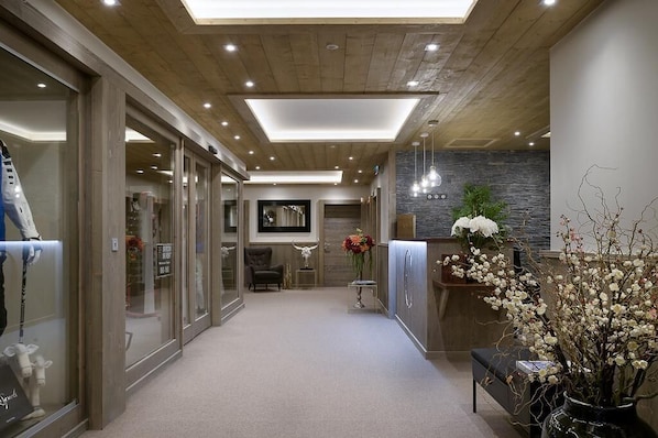 A modern hallway with a wooden ceiling, recessed lighting, a reception desk, and a seating area with a chair and a vase of flowers.