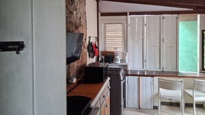 A kitchen with a stone wall, wooden beams, a TV, a blender, and a wicker chair. - Budget Adventurer's Hut (Bridgetown)