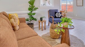 A spacious living room with a large brown sofa, a blue armchair, and a wooden coffee table. There are several potted plants and a small side table. - Stay Near Gulfstream, Outlets & Airport (Pooler)