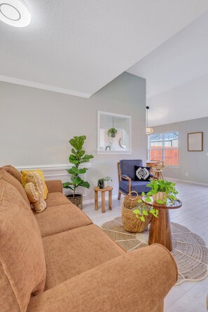 A spacious living room with a large sofa, a blue armchair, and a small table with a plant. There is a window with a view of a building outside.