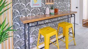 A room with a patterned wallpaper, a wooden bar counter, two yellow stools, and a framed picture on the wall. - Stay Near Gulfstream, Outlets & Airport (Pooler)