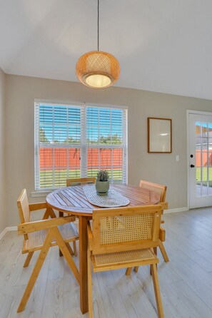 A dining area with a round wooden table, six chairs, a hanging light fixture, and a window with blinds.