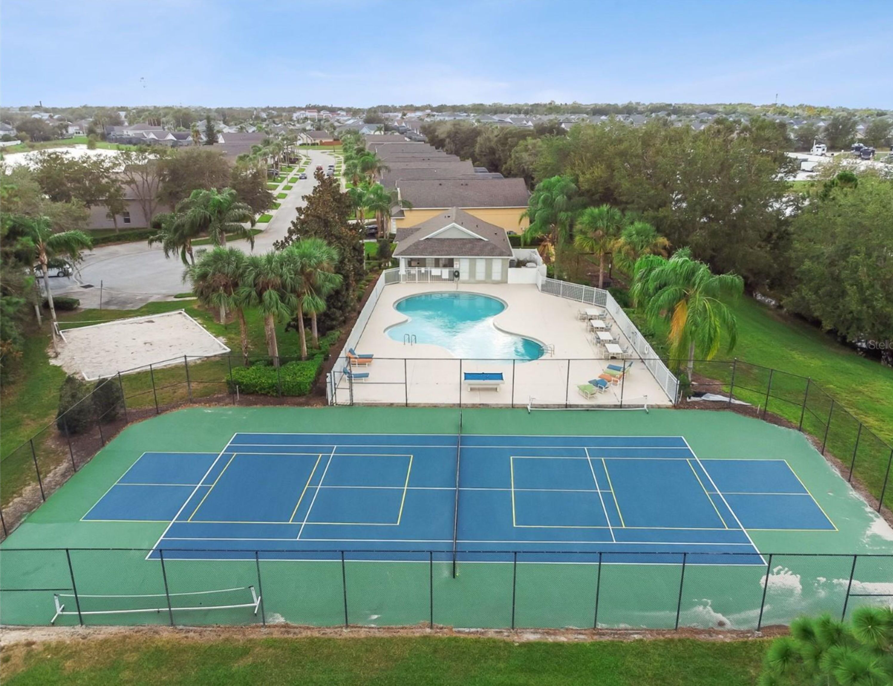 Una cancha de tenis con piscina y sillones de descanso, rodeada de palmeras y un área residencial al fondo.