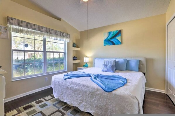 A bedroom with a large window, a bed with a blue blanket, a nightstand, a lamp, and a wall art piece. - Flounder Vacation Home (Clermont)