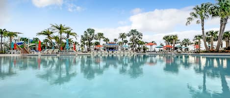 A large, clear swimming pool with lounge chairs and umbrellas, surrounded by palm trees and a building with a sign.