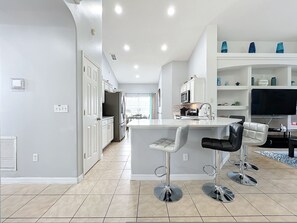 A modern kitchen with a central island, bar stools, a dining area with a TV, and a living space with shelves. - Beach Theme Vacation Pool Spa Home Mins to Disney (Clermont)