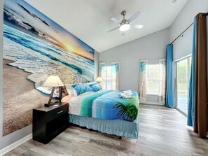 A bedroom with a large beach mural, a bed with a blue and green bedspread, two bedside lamps, a ceiling fan, and a large window with curtains. - Beach Theme Vacation Pool Spa Home Mins to Disney (Clermont)