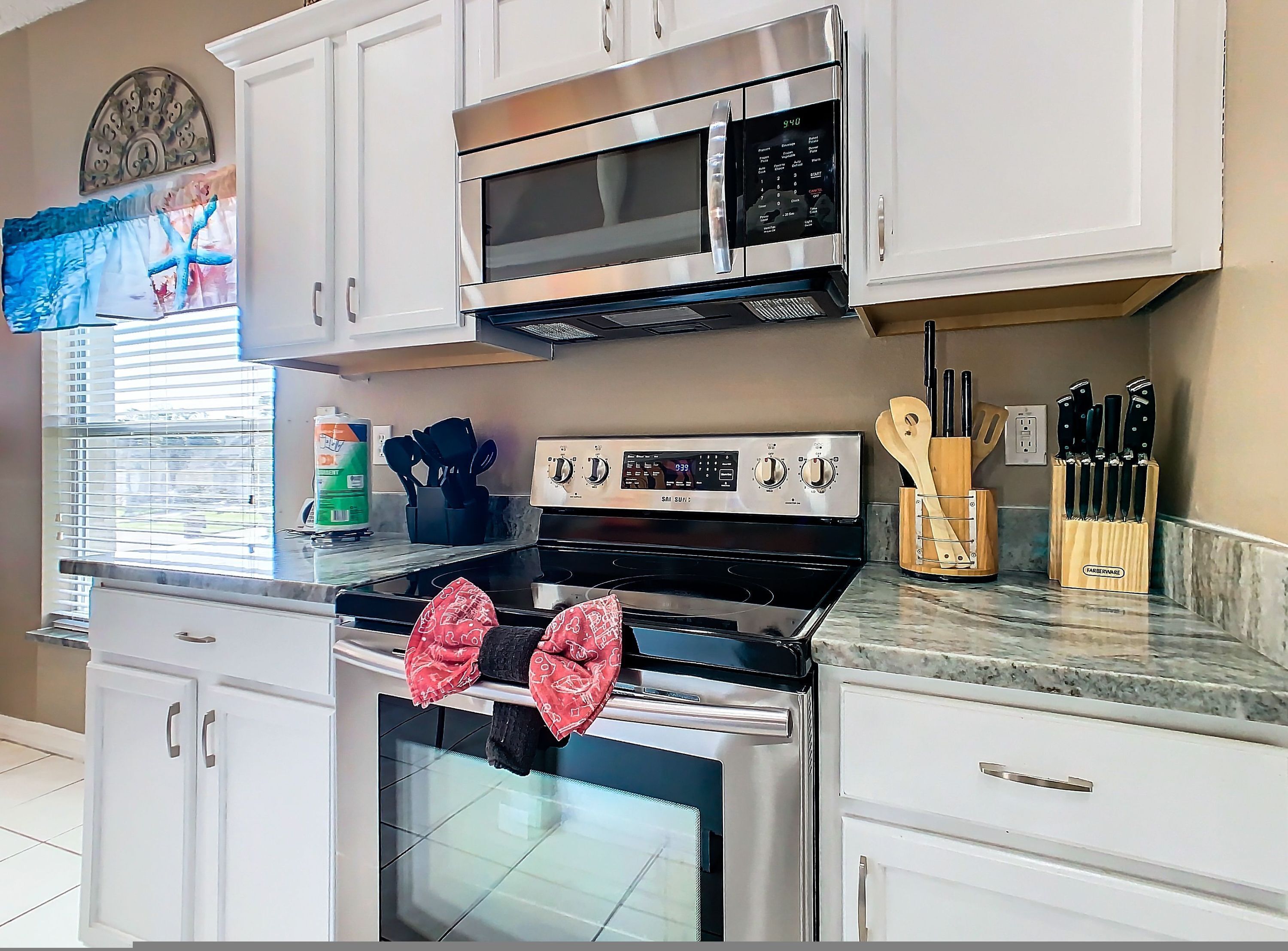 A kitchen with white cabinets, a stainless steel stove, a microwave, and a window with blinds.