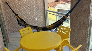 Een balkon met een gele tafel en stoelen, een hangmat en uitzicht op gebouwen.