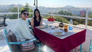 Two people seated at a table with a red tablecloth, a green umbrella, and a city view in the background. - Soham Haveli Udaipur (Udaipur)