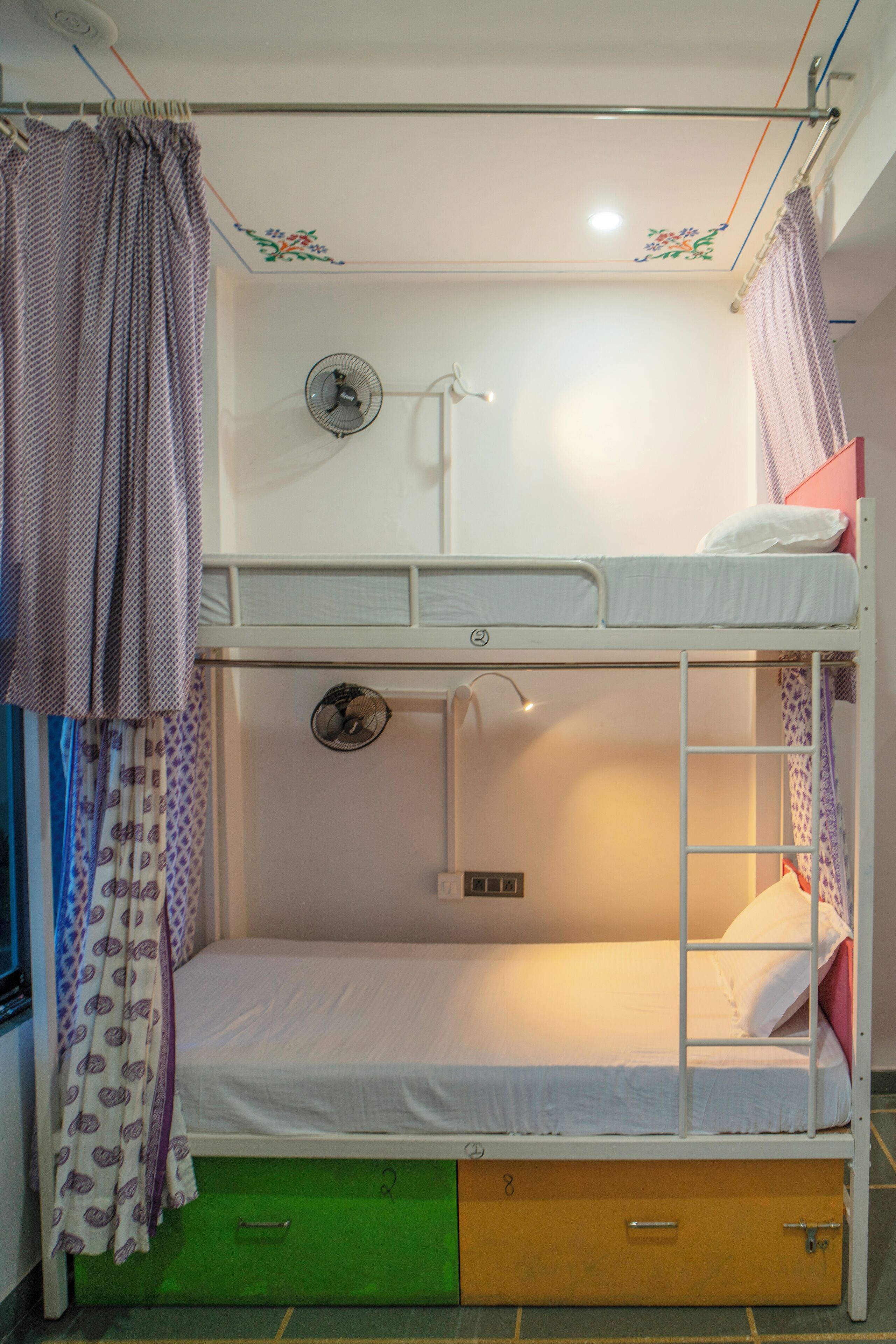 Una cama litera con un cajón verde, un ventilador pequeño y una ventana con cortinas estampadas.