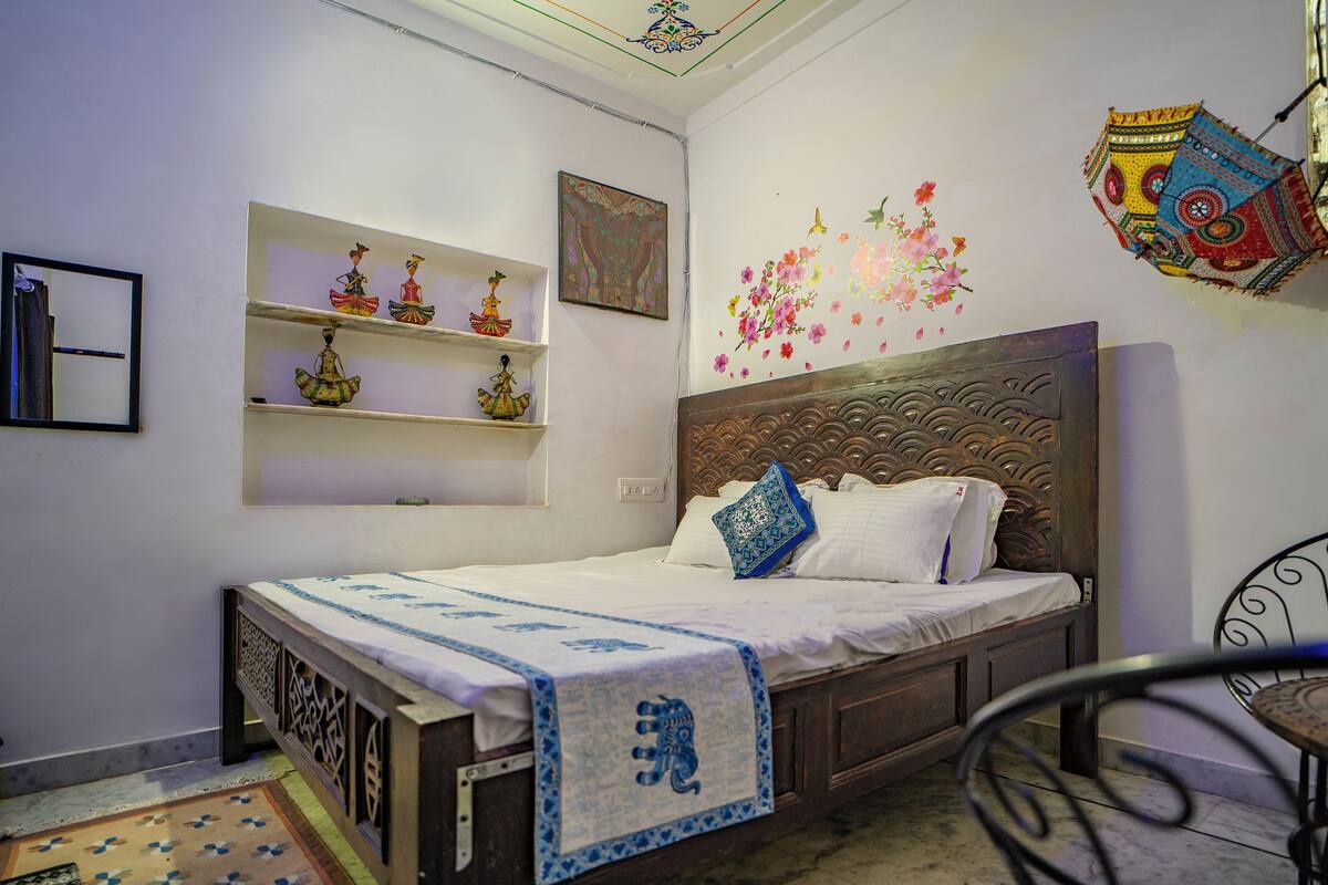 A bedroom with a wooden bed, white bedding, decorative pillows, a wall-mounted shelf with figurines, and a colorful hanging lantern.