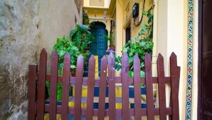 A narrow alleyway with a wooden gate leading to a building with a blue and yellow patterned mat at the entrance. - Soham Haveli Udaipur (Udaipur)