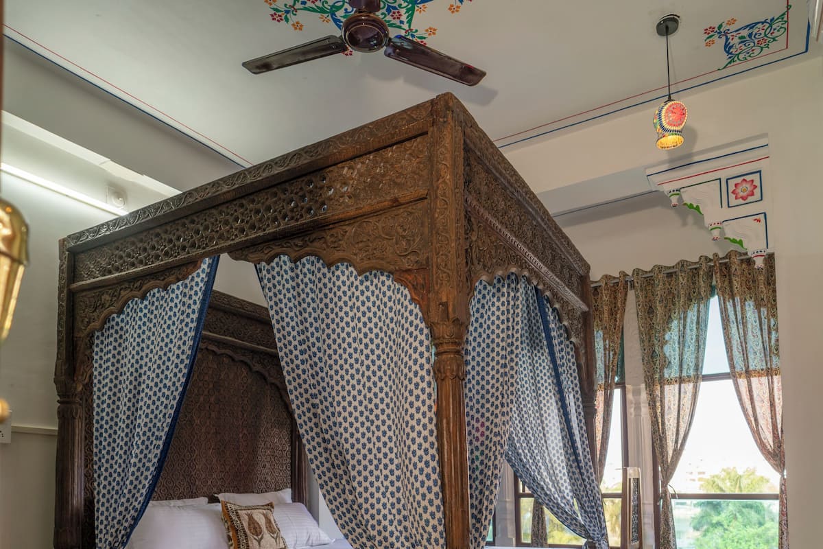 A four-poster bed with a canopy, a wooden ceiling fan, and a view of greenery outside.