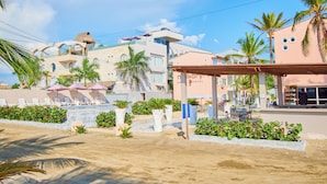 Um hotel à beira-mar com vários edifícios, palmeiras e uma área de areia com espreguiçadeiras.