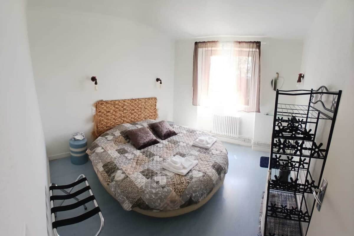A bedroom with a round bed, a wicker headboard, a radiator, a window with curtains, a black metal rack, and a striped chair.