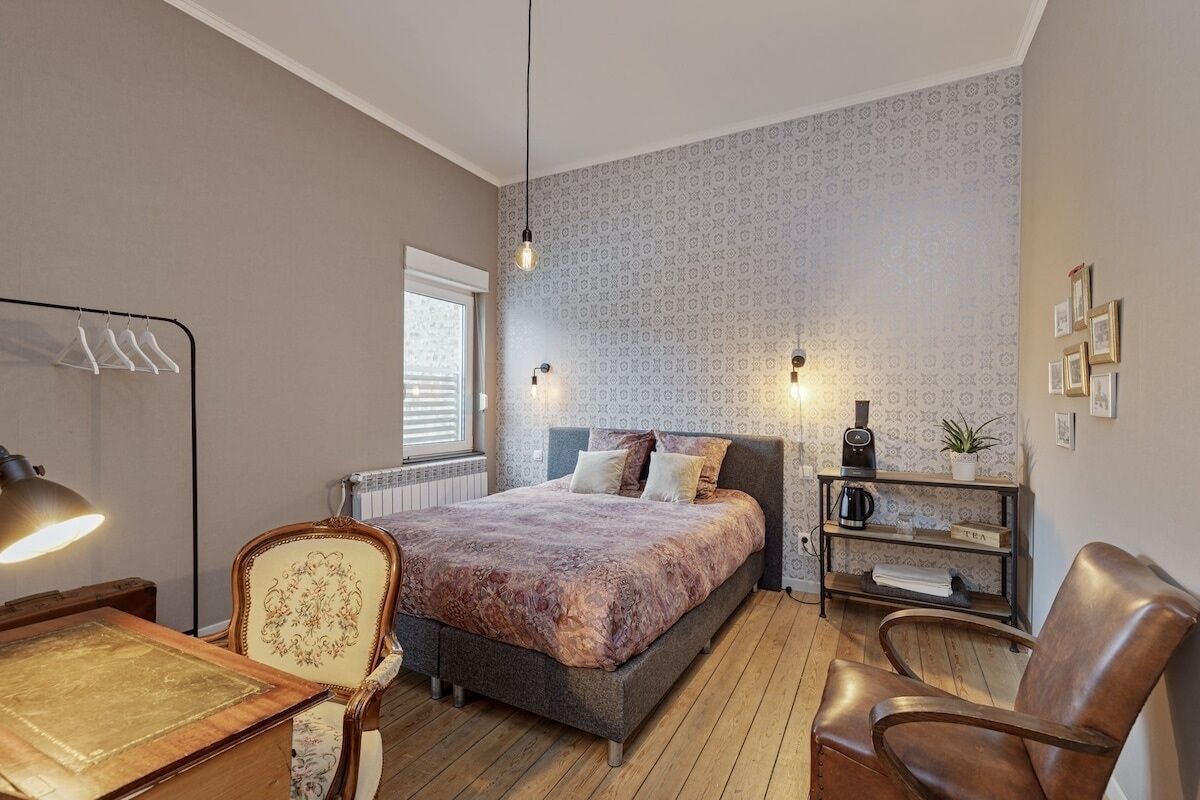 A bedroom with a bed, a desk, a chair, a radiator, a shelf with a plant, and a wall with framed pictures.