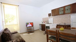 Un espace de vie compact comprenant un canapé, un téléviseur, une table à manger avec des chaises, un coin cuisine avec des placards et une fenêtre avec des stores.