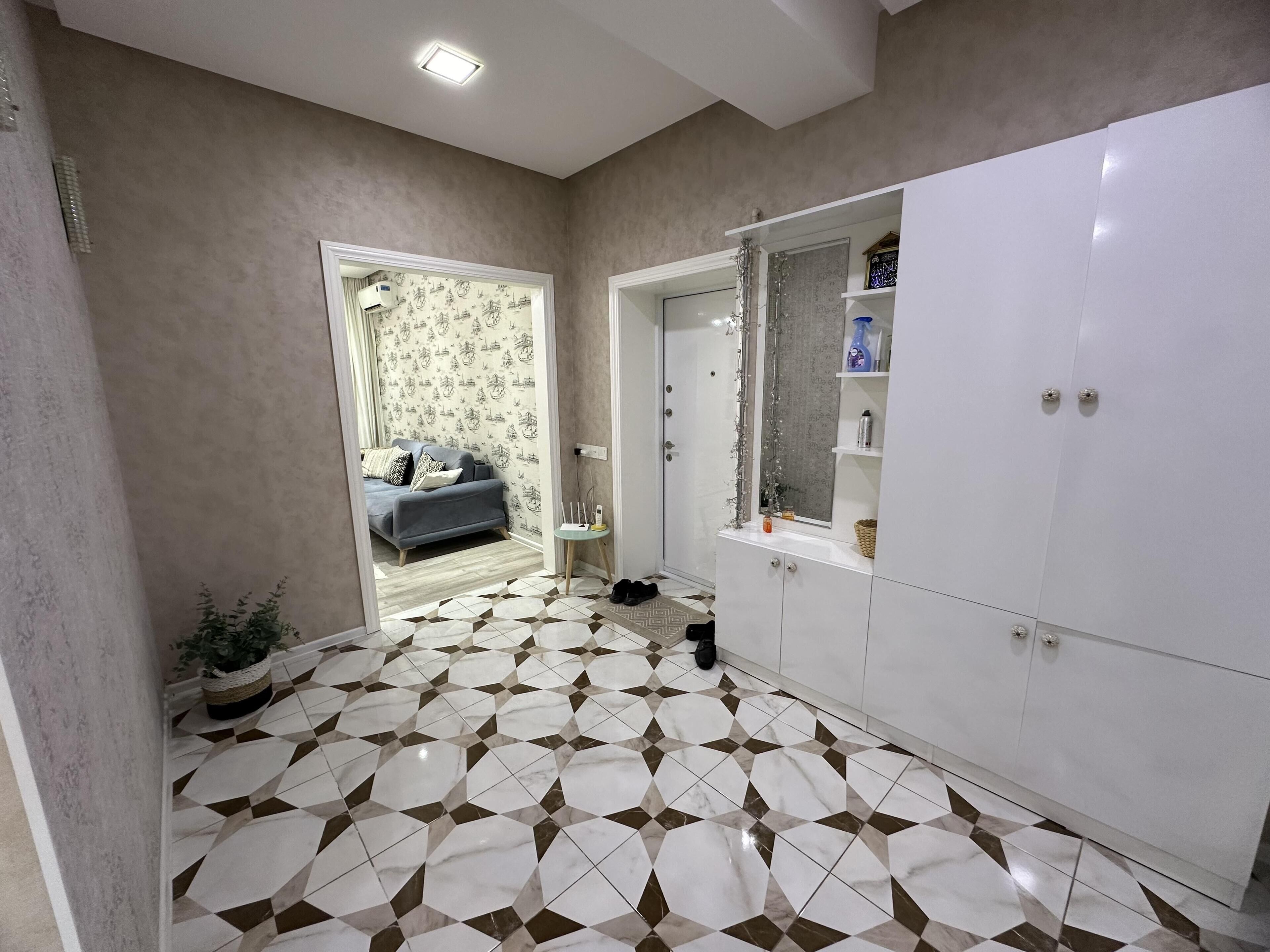 A room with a patterned floor, a grey sofa, a white wardrobe, and a bathroom with a shower and a mirror.