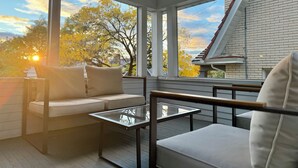 A sunlit balcony with a sofa and a glass table, overlooking a brick building and trees. - The Colonial Mt. Lebanon by Luxe PGH (Pittsburgh)