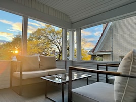 A sunlit balcony with a sofa and a glass table, overlooking a brick building and trees.