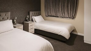 A bedroom with two beds, a nightstand with drawers, a lamp, and a large window with curtains.
