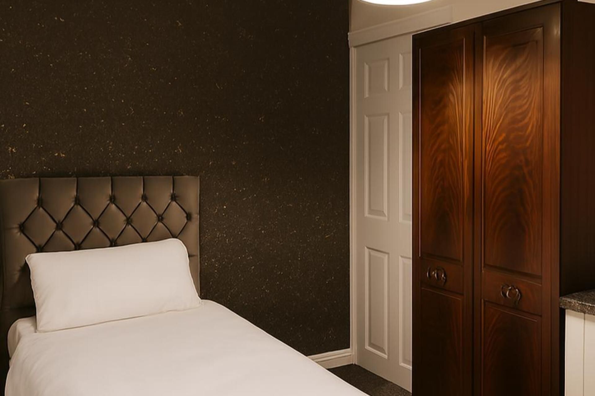 Un dormitorio con cabecera capitonada, cama blanca y pared oscura con textura.