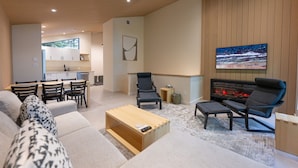 Un salon moderne avec un canapé, des fauteuils, une table basse et une cheminée. On aperçoit une cuisine à l’arrière-plan, avec des placards et une fenêtre.