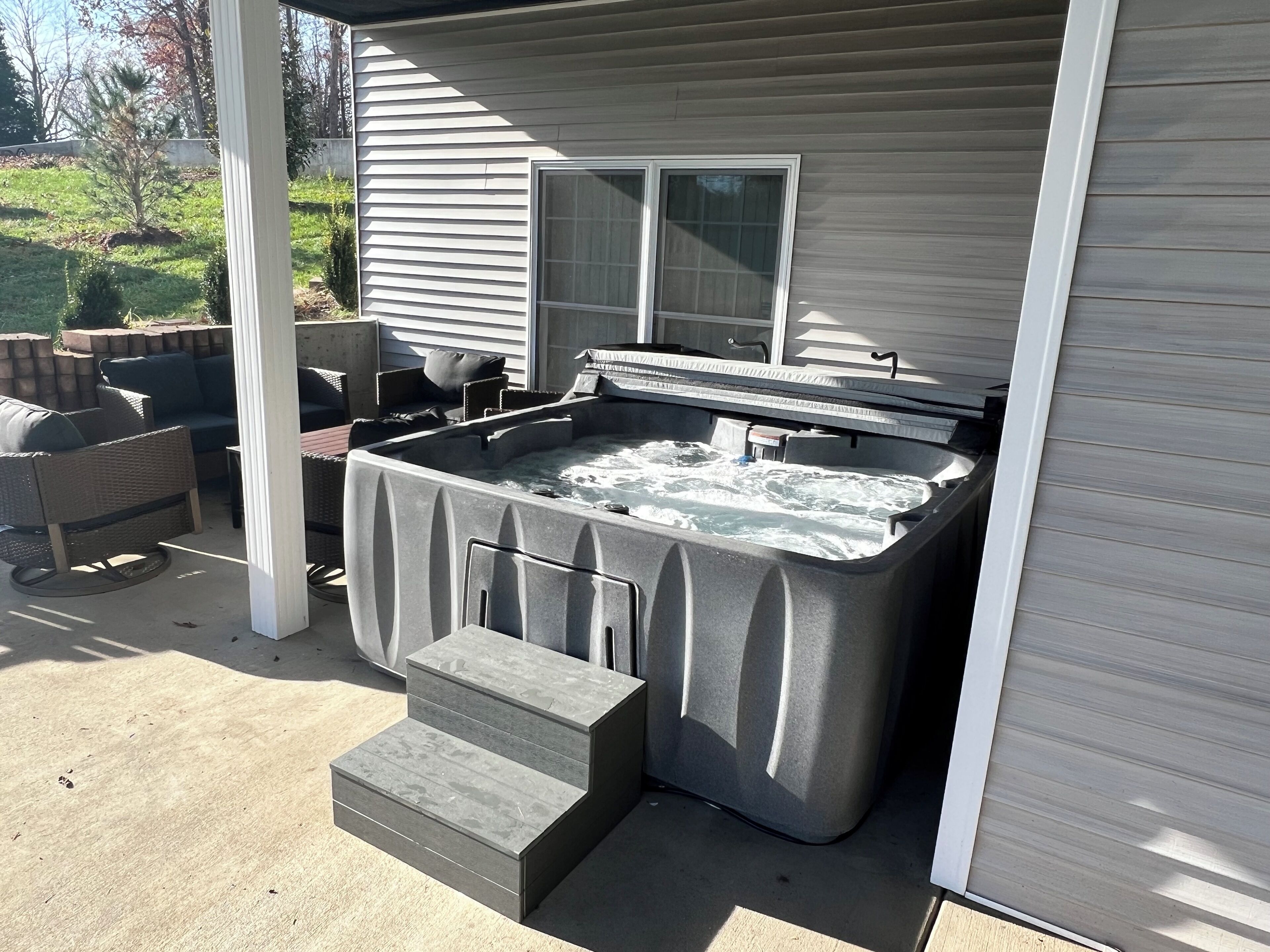 A hot tub with steps leading up to it, positioned under a covered patio area with outdoor furniture.