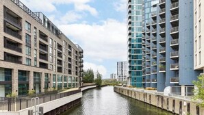 Un canal bordé d’immeubles modernes à plusieurs étages, dotés de balcons, un pont piétonnier et un ciel dégagé.