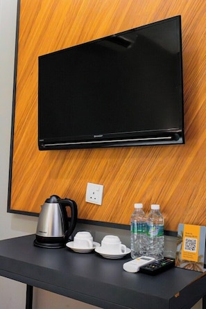 Ein Hotelzimmer mit Flachbildfernseher an einer Holzwand, einer Kaffeemaschine, Tassen, Wasserflaschen, einer Fernbedienung und einem QR-Code für den WLAN-Zugang.