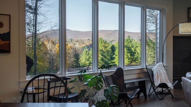 A room with large windows offering a view of a forested hillside. There are several chairs and a table in the room. - Spacious 6BR VT Retreat w/Sauna & Golf Simulator Near Stowe, Smuggs & Jay Peak (Johnson)