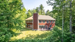 A two-story house with a red brick exterior, large windows, and a deck with outdoor furniture. - Spacious 6BR VT Retreat w/Sauna & Golf Simulator Near Stowe, Smuggs & Jay Peak (Johnson)